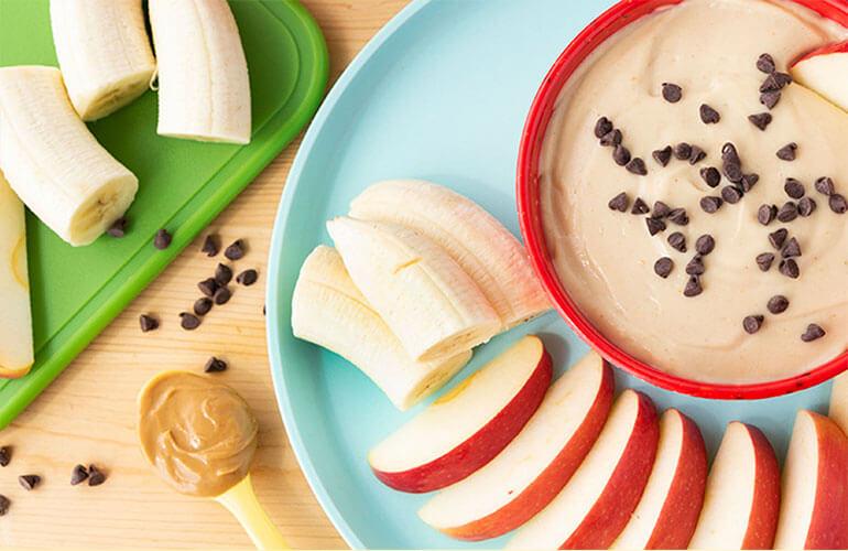 Bowl of peanut butter dip sprinkled with mini chocolate chips on a plate with sliced bananas and apples. A notebook with math homework and a pencil are nearby.