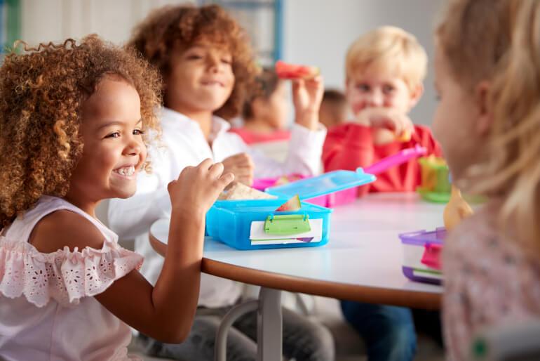 Small children sitting at a table eating lunch from their lunch boxes, smiling at each other.